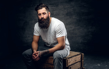 Bearded male with tattoo on arm dressed in a white t shirt sits on a wooden box.