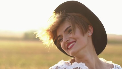 The emotions of the happy woman with red lipstick, cateyes and black vintage hat possing to the camera at the background of the field during the sunset.