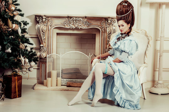 Indoors shot in the Marie Antoinette style. A young luxurious girl in a lush blue retro dress with a high hairstyle sits in a chair. Woman tired of luxury
