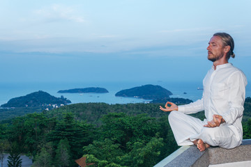 Yoga man sitting on rock fence. Male wearing white clothing sitting in lotus pose on stone fence with sea and mountains on background.