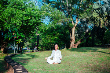 Man  asana on meadow. Young male in white clothing sitting in asana on green meadow in tropical park.