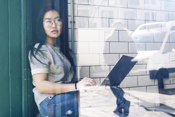 International student girl is studying online while sitting in a coffe shop with a laptop connected to the internet. Asian entrepreneur is typing e-mails to the partners by using a portable computer.