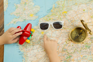Dreaming about summer holiday trip. Kid hand pointing to the map. Sun glasses, compass and toy red plane.  Top view 