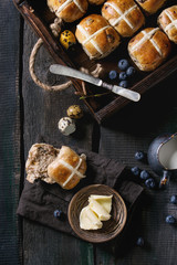 Hot cross buns in wooden tray served with butter, knife, blueberries, easter eggs, birch branch, jug of cream on textile napkin over old texture wood background. Top view, space. Easter baking.