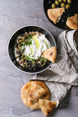 labneh middle eastern lebanese cream cheese dip with olive oil, salt, herbs, olives tapenade served in black bowl with traditional pita bread over gray texture metal background. Top view with space