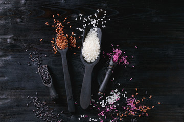 Variety assortment of raw uncooked colorful rice white, black, brown, pink in black spoons and scoop over burnt wooden background. Top view with space