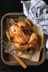 Grilled baked whole organic chicken on backing paper in old oven tray with white kitchen towel and meat fork over black burnt wooden background. Top view with space.