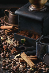Black roasted coffee beans and grind with spices cinnamon, anise, cardamom, clove and brown sugar. With black vintage coffee grinder, scoops and terracota cup over wood burnt background.