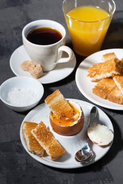 soft boiled egg, toasts, cup of coffeeand orange juice for breakfast on dark background, vertical