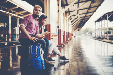 Young tourist couple with backpack sitting on bench at the train station. Two young tourist are waiting to get on the train and begin their journey. Travel concept.