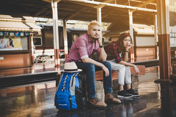 Obraz na płótnie Canvas Hipster couple sitting on bench at the train station. Two young tourist are waiting to get on the train and begin their journey. Travel concept.