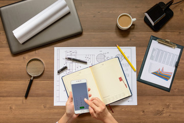 Girl is sitting at table with business accessories, cup of coffee and a laptop, works with drawings, graphs, tables, using smartphone. Top view.