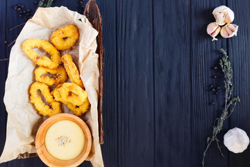 Fried onion rings in breadcrumbs served with creamy sauce, herbs and spices on craft paper and bark, free space for text.