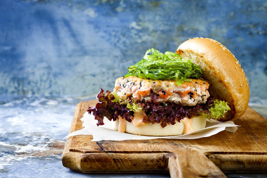 Asian style salmon burger with grilled shrimps, seaweed, lettuce and spicy sriracha mayo sauce served on pieces of brown paper on a rustic wooden board. Bright blue background with copyspace