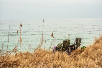 Fototapeta na wymiar Folding chairs and a couple sitting on the seashore. Calm sea view. The shore is covered with dry grass. Early spring. 