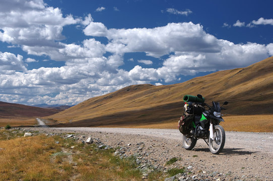 Motorcycle traveler enduro with suitcases standing on a dirt road vanishing into the skyline under a blue sky with white clouds, Plateau Ukok, Altai mountains, Siberia, Russia.