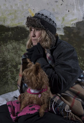Mature homeless woman with a Yorkshire terrier leaning against an old wall 