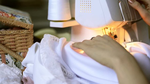 Seamstress sews satin fabric on overlock inside atelier. Close up view on hands of tailor, young woman, with beige nails and rings on fingers, professionally and quickly stitches edge of white, laced