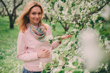 happy pregnant blonde beautiful woman walking outddor in spring park or garden, posing near blooming tree