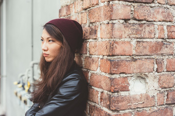 Portrait of asian beautiful young woman outdoor in the city posing leaning on a brick wall looking away serious - indipendent, girl power, confidence concept