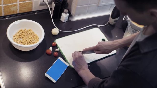 Hands of man cutting garlic at home kitchen and watching video at smartphone
