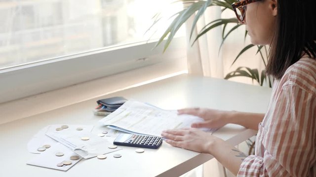 Young woman in glasses lives alone in small apartment and tries calculate her household bills, desperately shakes her head while looking at paper