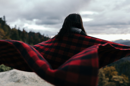 Person covered in blanket looking at view of mountains, woodland and stormy sky 