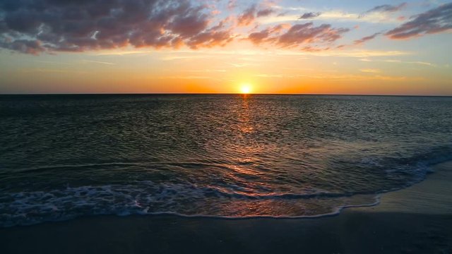 The setting sun paint the sky with color as shorebirds scurry about in the gentle surf breaking on a sandy Florida beach in this looping video footage.
