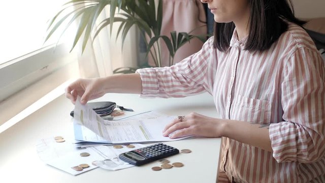 Young brunette woman in striped shirt sits at table next to window and counts her household bills looking at numbers in papers and using scientific calculator to check total sum.