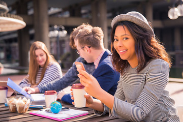 Fototapeta premium Happy students sit in time of a break and discuss about their tasks, with coffee, with a phones, with the folders, with the foods and one of them keeps a phone and cup of coffee in her hands, outdoors