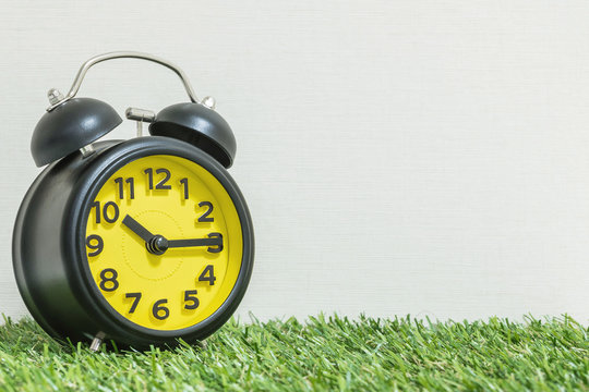 Closeup black and yellow alarm clock for decorate show a quarter past ten o'clock or 10:15 a.m. on green artificial grass floor and cream wallpaper textured background with copy space
