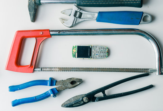 broken cellphone on white desk with wrong tools for repair. hammer, hacksaw, pliers, wrench and metal shears. fun concept