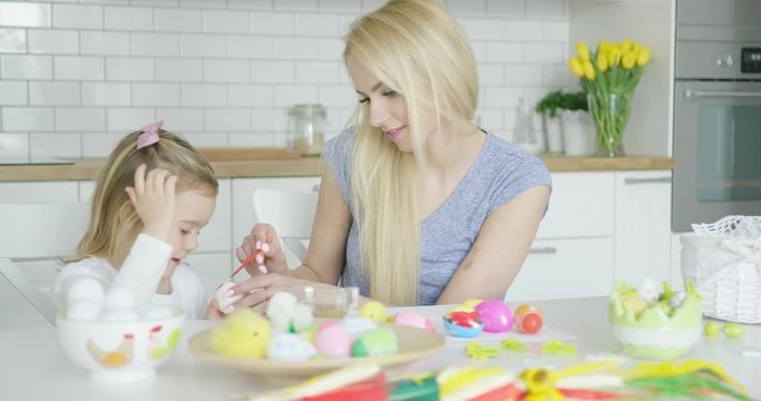 Laughing young mother and little adorable girl coloring Easter eggs together while sitting at table in kitchen. 