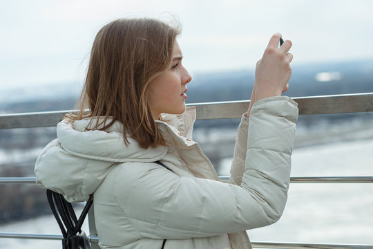 Young adorable student blonde teen girl takes photos with smartphone on the observation deck with a view of cloudy spring sky, frozen river, sunny windy weather