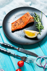 One piece of baked salmon with lemon on a black plate. Blue wood background