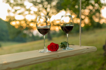 Two glasses of red wine and beautiful red rose lying on a swing