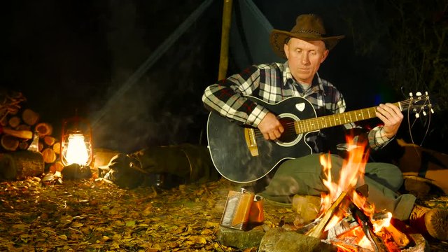 Adult man in the cowboy hat  playing guitar near the bonfire.