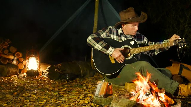 Adult man in cowboy hat sitting near the campfire and tuning guitar outdoors.