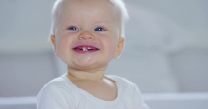 A baby, a boy with large blue eyes and light-colored hair, sits and smiles on a snow-white blanket, looks at her mother, on a white background.