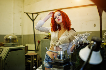 Fototapeta na wymiar Red haired girl wear on short denim shorts and white blouse posed at industrial machine at the factory.