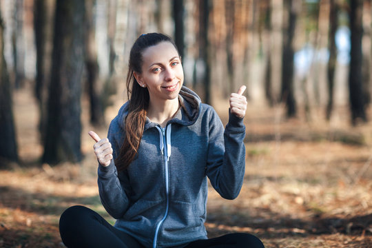Young woman sitting on the grass in the forest on sunny day, shows thumb up