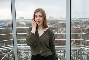 Young adorable student blonde teen girl touching hair showing tongue having fun on the observation deck with a view of cloudy spring sky, frozen river, sunny windy weather