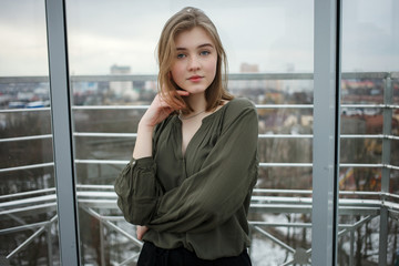 Young adorable student blonde teen girl touching hair having fun on the observation deck with a view of cloudy spring sky, frozen river, sunny windy weather