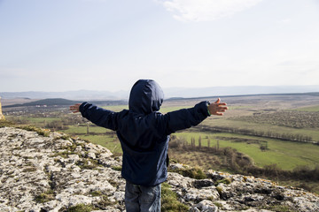 A little boy in a blue jacket is standing on a mountain. White cliff (Ak-Kaya) in the Crimea. Stone blocks and views of the wide steppe. Spring, green grass and clear sky