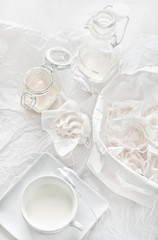 High key still life with meringues, crumbs, glass jars and cup with milk and paper roses.