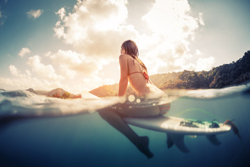 Split shot of the young woman sitting on the surfboard in the ocean with green sunrise hills on the...