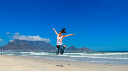 Beautiful girl jumping at Milnerton beach, Cape Town. Table Mountain and breaking waves in the background. Concept photo of happiness, freedom and lifestyle.