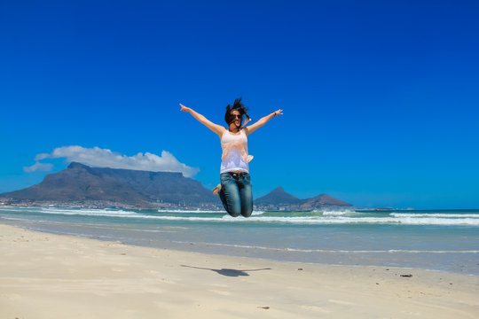 Beautiful girl jumping at Milnerton beach, Cape Town. Table Mountain and breaking waves in the background. Concept photo of happiness, freedom and lifestyle.