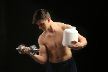 Young handsome sportsman holding bottle of protein and dumbbell on black background