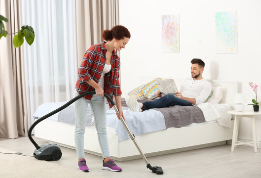 Man reading newspaper on bed while woman hoovering floor at home